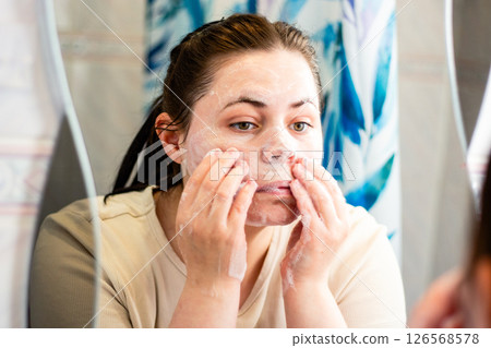 Young woman washing face with foaming cleanser in bathroom mirror. Perfect for skincare routines beauty tutorials. International self care day. Young woman washing face with foaming cleanser in bathroom mirror. Perfect for skincare routines beauty tutorials. International self care day. 126568578