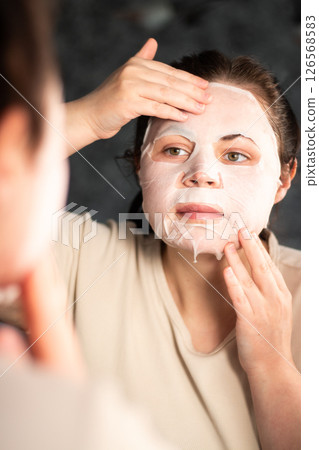 Close up of female adjusting sheet mask on face in modern bathroom. Ideal for cosmetic ads or spa treatment visuals. 126568583