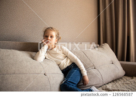Young girl with blonde hair, sitting casually on soft gray couch, resting her chin on her hand, in cozy living room setting, showcasing relaxed home atmosphere 126568587