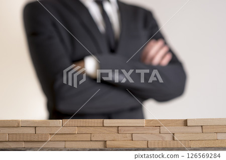 Businessman with folded arms with wall of wooden bricks infront of him Businessman with folded arms with wall of wooden bricks infront of him 126569284