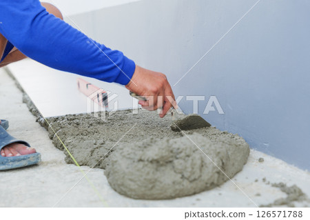 A worker hand uses a trowel to smooth gray mortar, preparing a level surface for floor tiles. Precision and craftsmanship in tiling work are on display. 126571788