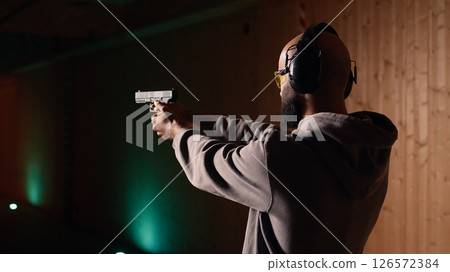 Shooting range customer shooting targets with pistol, learning to operate handgun in close quarters. Man at indoor shooting ground firing lethal fire weapon, wearing protection equipment, camera A Shooting range customer shooting targets with pistol, learning to operate handgun in close quarters. Man at indoor shooting ground firing lethal fire weapon, wearing protection equipment, camera A 126572384