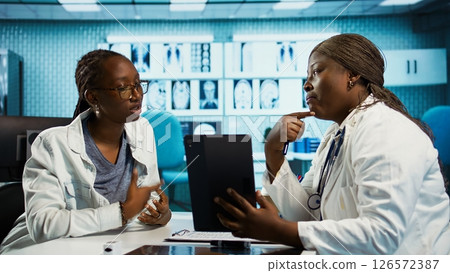 Black specialist providing checkup examination on insurance with patient, employing technology for diagnostic tools in clinic. Girl asking physician about wellness advice, recovery plan. Camera B. Black specialist providing checkup examination on insurance with patient, employing technology for diagnostic tools in clinic. Girl asking physician about wellness advice, recovery plan. Camera B. 126572387