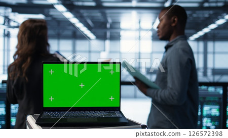 Green screen laptop in front of data center engineers in blurry background calibrating energy usage, inspecting servers configuration, supporting sustainable infrastructure, camera B 126572398