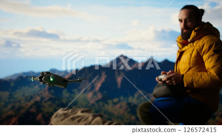 Traveler balances adventure and creating content by filming with his drone, recording the aerial view of a beautiful mountain range. Breathtaking landscape scenery videography. Camera A. 126572415