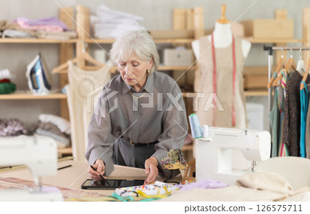 Mature female dressmaker drawing in tablet in sewing workshop Mature female dressmaker drawing in tablet in sewing workshop 126575711