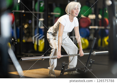 Mature woman trains muscles of arms, standing up doing barbell exercises 126575788
