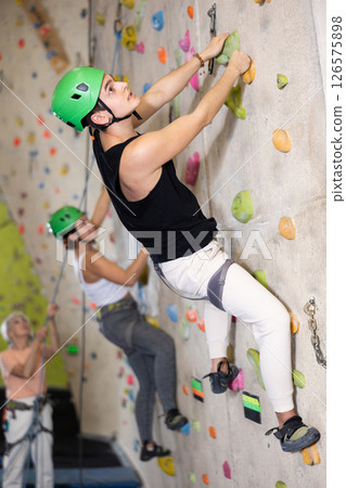Sporty young guy in safety gear doing a difficult wall climb in climbing gym Sporty young guy in safety gear doing a difficult wall climb in climbing gym 126575898