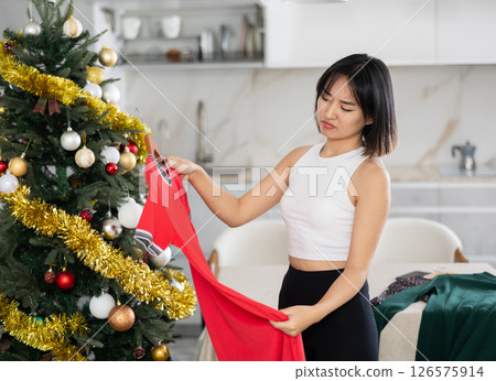 Sad young Asian girl trying to choose a dress standing next to Xmas tree 126575914
