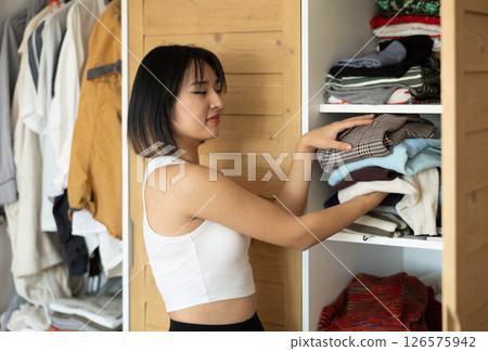 Happy young Asian girl putting some items of clothing in the wardrobe Happy young Asian girl putting some items of clothing in the wardrobe 126575942