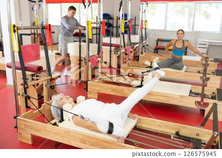 Elderly woman is engaged in Pilates simulator with cable and ropes for stretching leg muscles Elderly woman is engaged in Pilates simulator with cable and ropes for stretching leg muscles 126575945