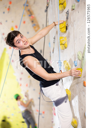 young positive man, insured by trainer, moving up artificial climbing wall indoors young positive man, insured by trainer, moving up artificial climbing wall indoors 126575965