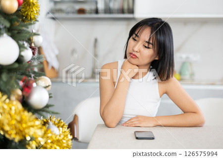 Sad young Asian girl waiting for phone call sitting at table by Xmas tree Sad young Asian girl waiting for phone call sitting at table by Xmas tree 126575994