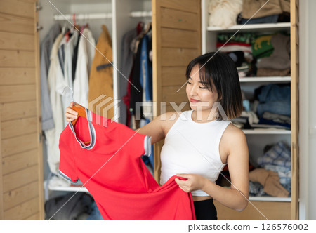 Happy young Asian girl trying to choose a dress standing by wardrobe Happy young Asian girl trying to choose a dress standing by wardrobe 126576002