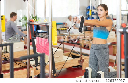 Sports club girl client on reformer with cable and rope performs exercise to stretch muscles of arms Sports club girl client on reformer with cable and rope performs exercise to stretch muscles of arms 126576017