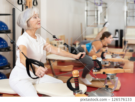 Sports club senior woman client performs exercise on reformer with cable and rope for arms 126576133