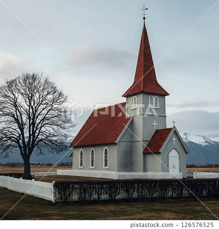 A small grey church with a red roof and steeple stands beside a bare tree, surrounded by a white fence in a field 126576525