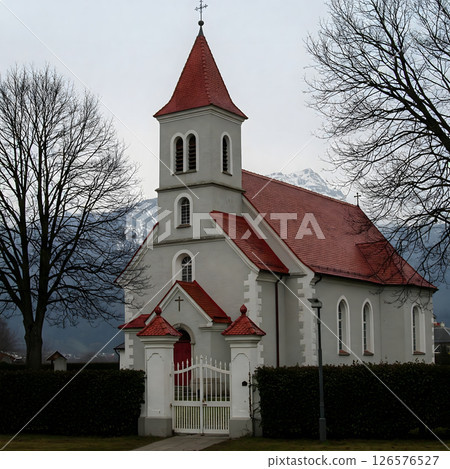 Grey church with a red roof and steeple, surrounded by bare trees and distant mountains 126576527