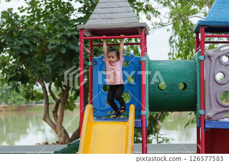happy toddler girl hanging the red bar by her hand at outdoor playground in park 126577583