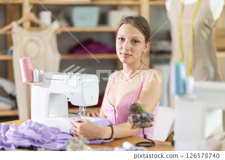 Positive young female dressmaker working with sewing-machine in tailor's workshop 126578740