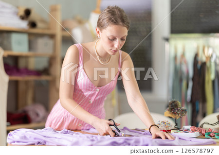 Young female dressmaker cutting textile in sewing workshop 126578797