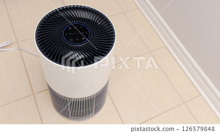 Tall round air purifier with a black top fan and a white body on a tile floor against the wall 126579848