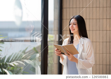 Smiling woman holding a notebook, looking out a window in a bright modern space 126580861
