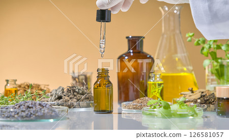 Close-up of essential oil being extracted from amber glass bottle with dropper in natural science research setup. White lab table with herbs displayed around it Close-up of essential oil being extracted from amber glass bottle with dropper in natural science research setup. White lab table with herbs displayed around it 126581057