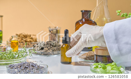 Scientist measuring yellow herbal extract into glass flask on white table, surrounded by dried plants and oil bottles.Herbal Research in the Laboratory 126581058