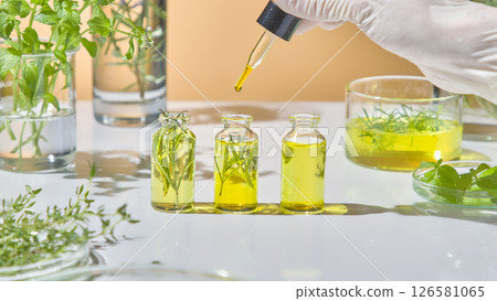 Filling small glass vials with herbal oil blend, arranged with natural ingredients in an organic skincare lab. Herbal leaves such as tea tree, rosemary... with yellow essential oil 126581065