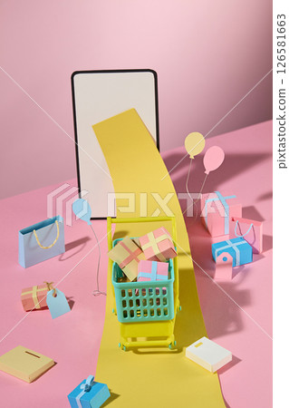 A long yellow receipt flows from the phone screen into the shopping cart, surrounded by colorful boxes, creatively displaying digital transactions. Colorful handmade props decorate the pink background 126581663