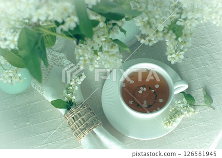 Cup of tea on the table with spring fresh bouquet with white blooming fragrant branches of bird cherry. Cup of tea on the table with spring fresh bouquet with white blooming fragrant branches of bird cherry. 126581945