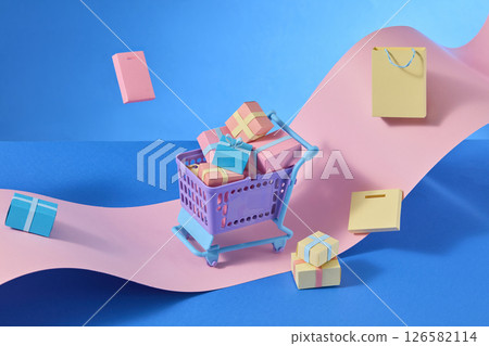Miniature shopping trolleys and pastel gift boxes with striking paper shopping bags, all arranged on abstract pink and blue paper waves symbolizing online shopping 126582114