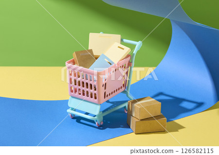 Bright pink mini shopping cart with colorful items placed on layered paper, evoking fun and whimsical elements of shopping and retail environments. Creative photo, shopping concept 126582115