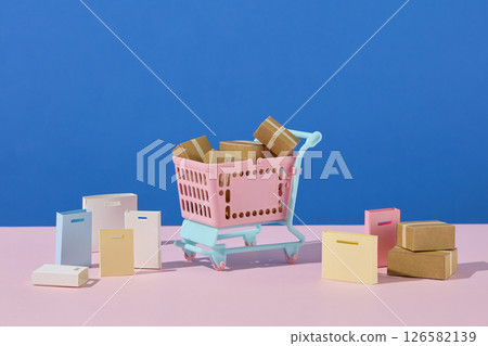 Pink cart filled with boxes and surrounded by small packages, arranged on a soft pink and blue background for a clean shopping-themed visual. Eye-catching color and empty space for design Pink cart filled with boxes and surrounded by small packages, arranged on a soft pink and blue background for a clean shopping-themed visual. Eye-catching color and empty space for design 126582139