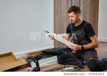 Man in overalls assembles furniture, examining white panel and surrounded by tools. Handyman in work clothes preparing parts pf cabinet to assembly. DIY furniture project 126583822