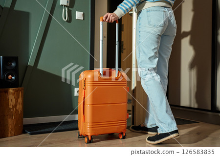 Person leaving home with orange suitcase. Female traveler start trip with luggage. Woman in casual outfit stands near door holding suitcase handle. Travel concept Person leaving home with orange suitcase. Female traveler start trip with luggage. Woman in casual outfit stands near door holding suitcase handle. Travel concept 126583835