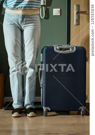 Close up of woman wearing jeans and shoes standing near suitcase ready to leaving home for travel. Female feet and luggage near door before departure 126583836