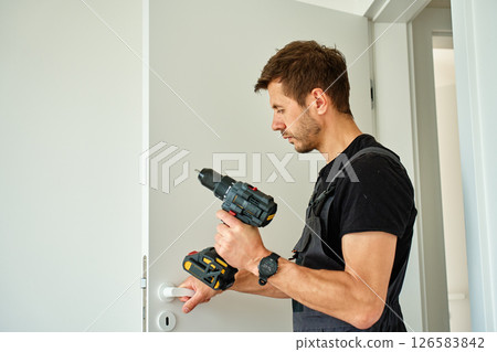 Man holds cordless drill and installs white door handle in modern interior. Handyman fitting door in apartment, using screwdriver. Concept of hardware installation during renovation 126583842