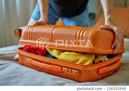 Woman trying to close suitcase at home. Female press down overfilled orange suitcase while packing clothes. Person preparing for departure Woman trying to close suitcase at home. Female press down overfilled orange suitcase while packing clothes. Person preparing for departure 126583848