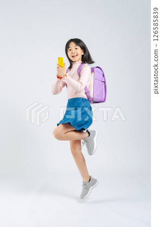 A full-body photo of an elementary school girl with a backpack jumping while using a smartphone 126585839