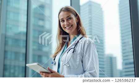 Confident young female doctor wearing a white lab coat and stethoscope, smiling while holding a digital tablet in a modern hospital or medical office with large windows. 126586237