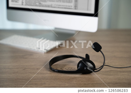 Call center black headset with microphone resting wooden desk front of computer monitor and keyboard, suggesting workspace or call center Call center black headset with microphone resting wooden desk front of computer monitor and keyboard, suggesting workspace or call center 126586283