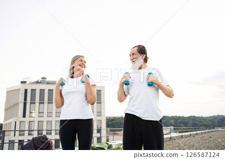 Elderly man and woman performing morning exercises with dumbbells outdoors on urban rooftop. 126587124