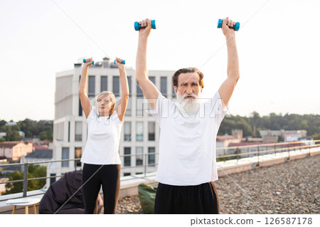 Elderly man and woman exercising with dumbbells outdoors, prioritizing health and fitness goals. Elderly man and woman exercising with dumbbells outdoors, prioritizing health and fitness goals. 126587178