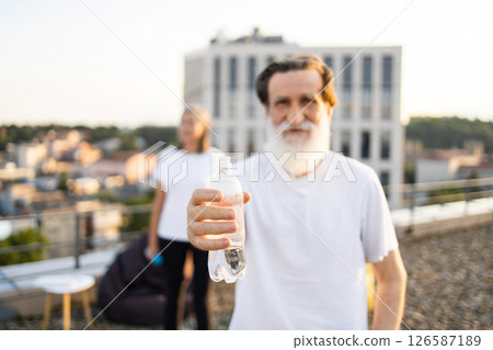 Elderly man with beard dressed in white shirt on urban rooftop holding water bottle. Background features a blurry cityscape. Scene exudes fitness, hydration, maturity, and enjoying active lifestyle. 126587189