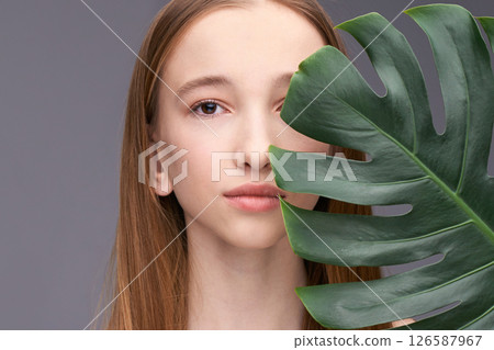 Young caucasian teenage female with long hair partially hidden behind monstera 126587967