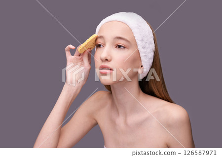 Young caucasian female with headband cleansing face using natural sponge Young caucasian female with headband cleansing face using natural sponge 126587971