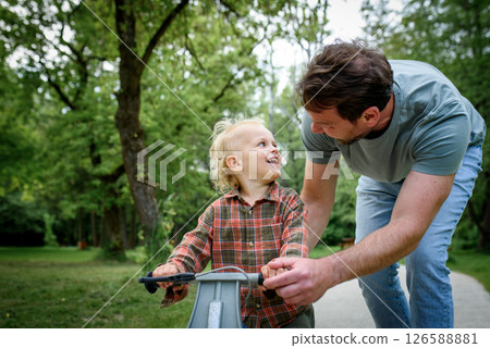 Proud dad watching son ride his balance bike in the park. Proud dad watching son ride his balance bike in the park. 126588881