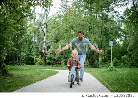Toddler riding balance bike with dad cheering behind 126588883